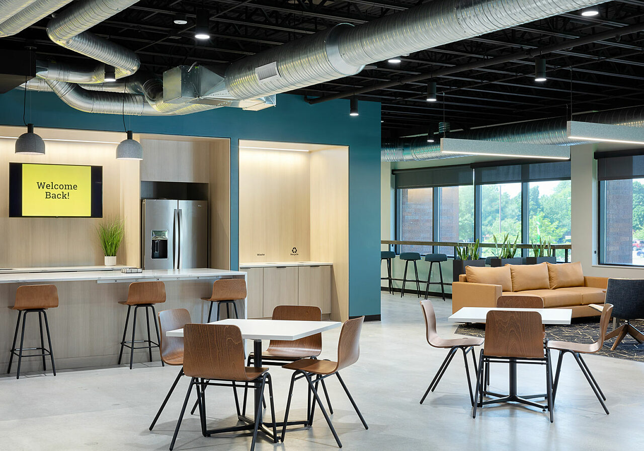 Watco breakroom features large counter with wood/metal stools, low white cafe tables with chairs that match stools. View of kitchen area, as well as couch.