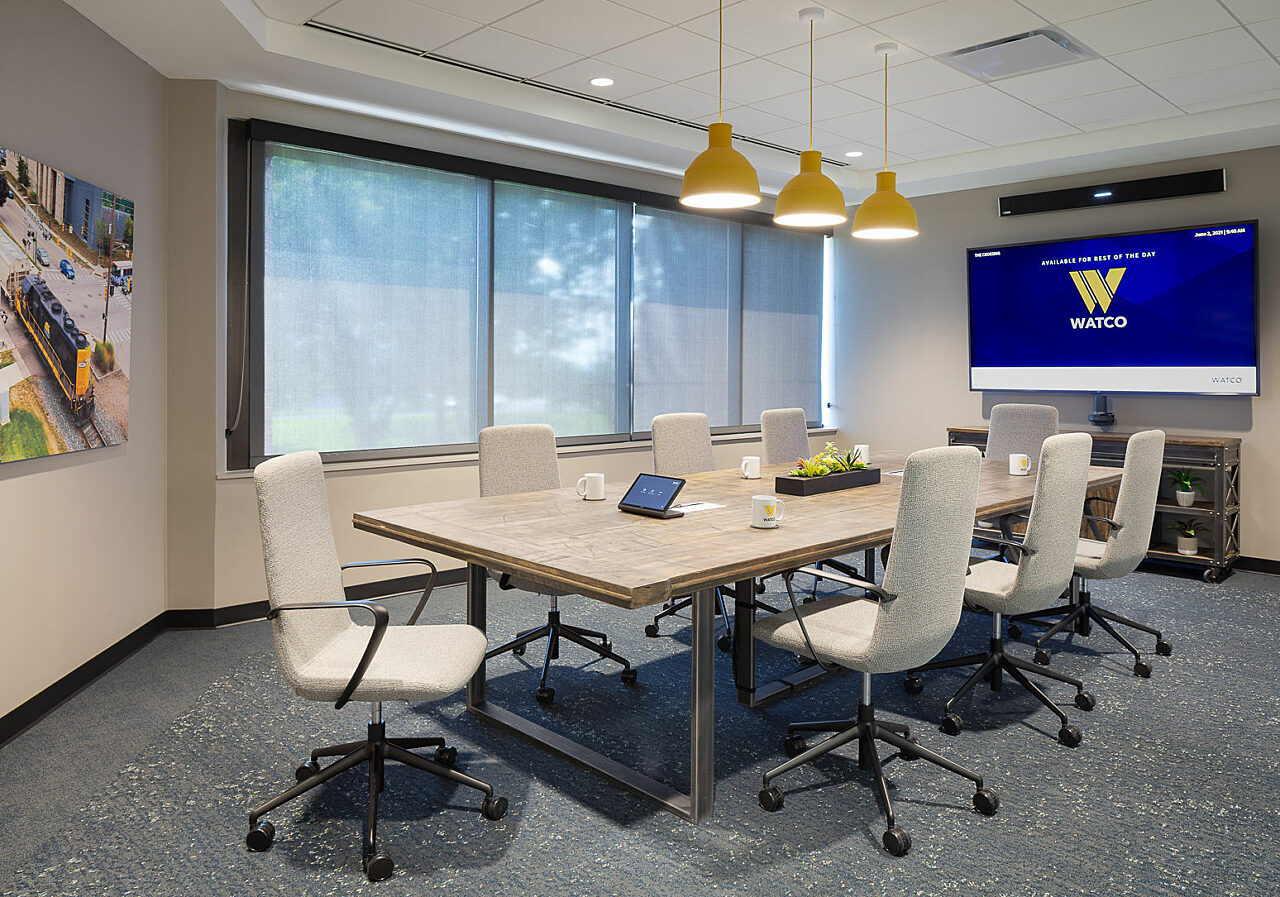 Watco executive conference room w/ custom table, off-white upholstered rolling chairs and yellow drop pendant lights. Railroad art and large screen w/ logo