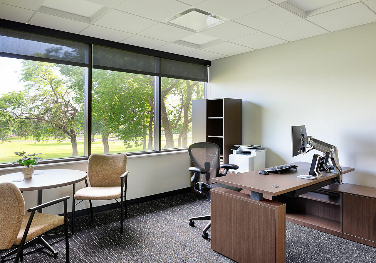 Watco private office with small meeting table and textured upholsetered chairs. Sit to stand desk and Aeron chair.  View of trees outside large windows.