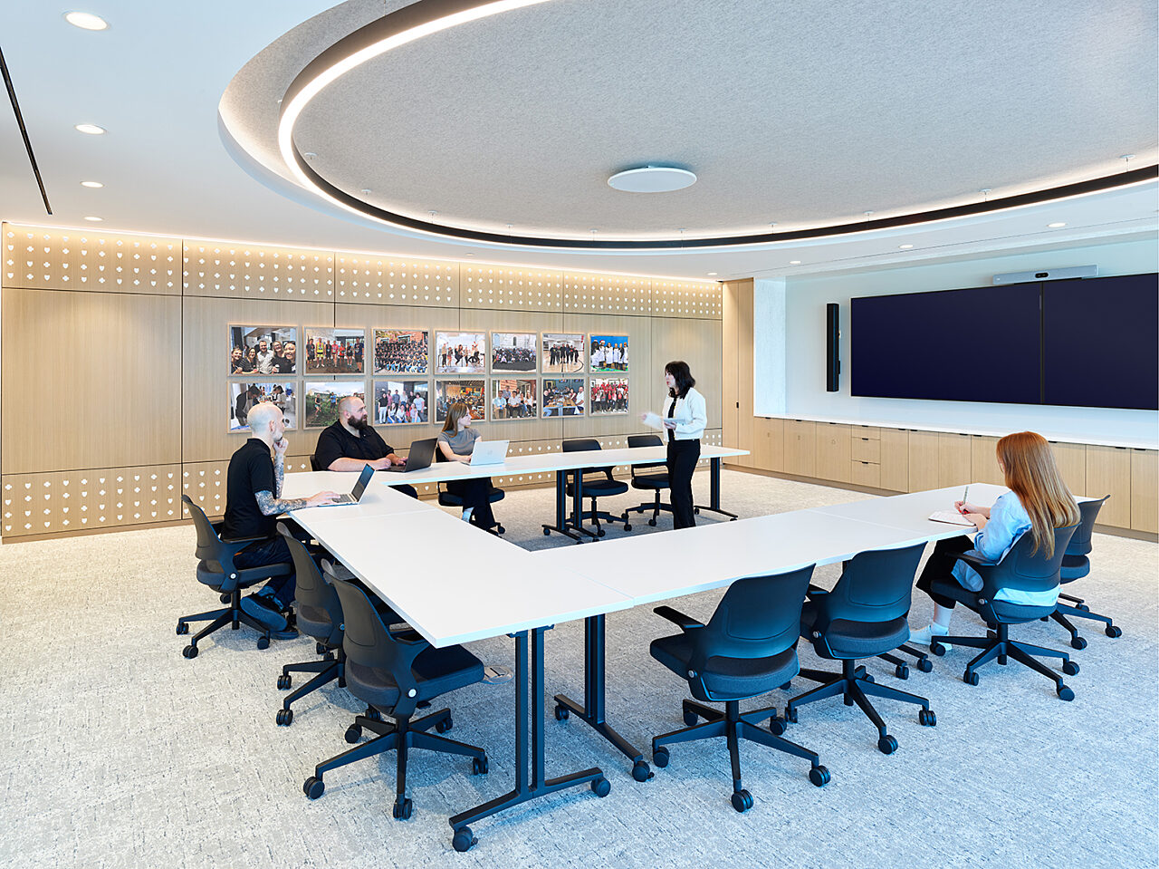 Employees interact in conference room with U-shaped table and blue/black chairs. Light wood wall features employee pics. Dual screens mounted on other wall
