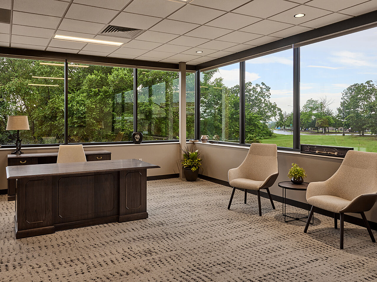 Tribal leader office with a large wooden desk. Two beige/wood lounge chairs with small table between. Continuous windows along two walls with tree views