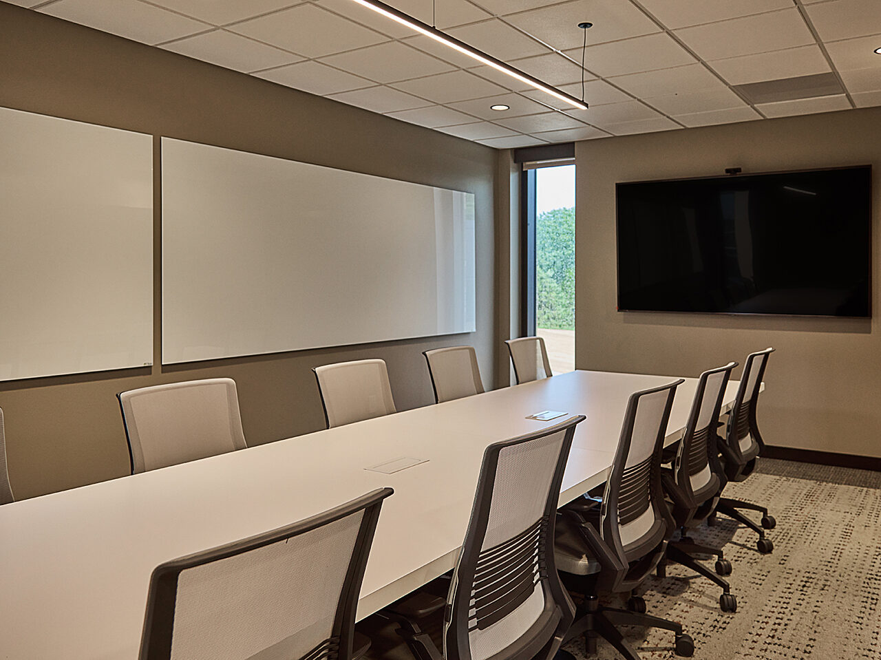 Conference room with white table and mesh-backed task chairs. Mounted TV and easel boards on walls.