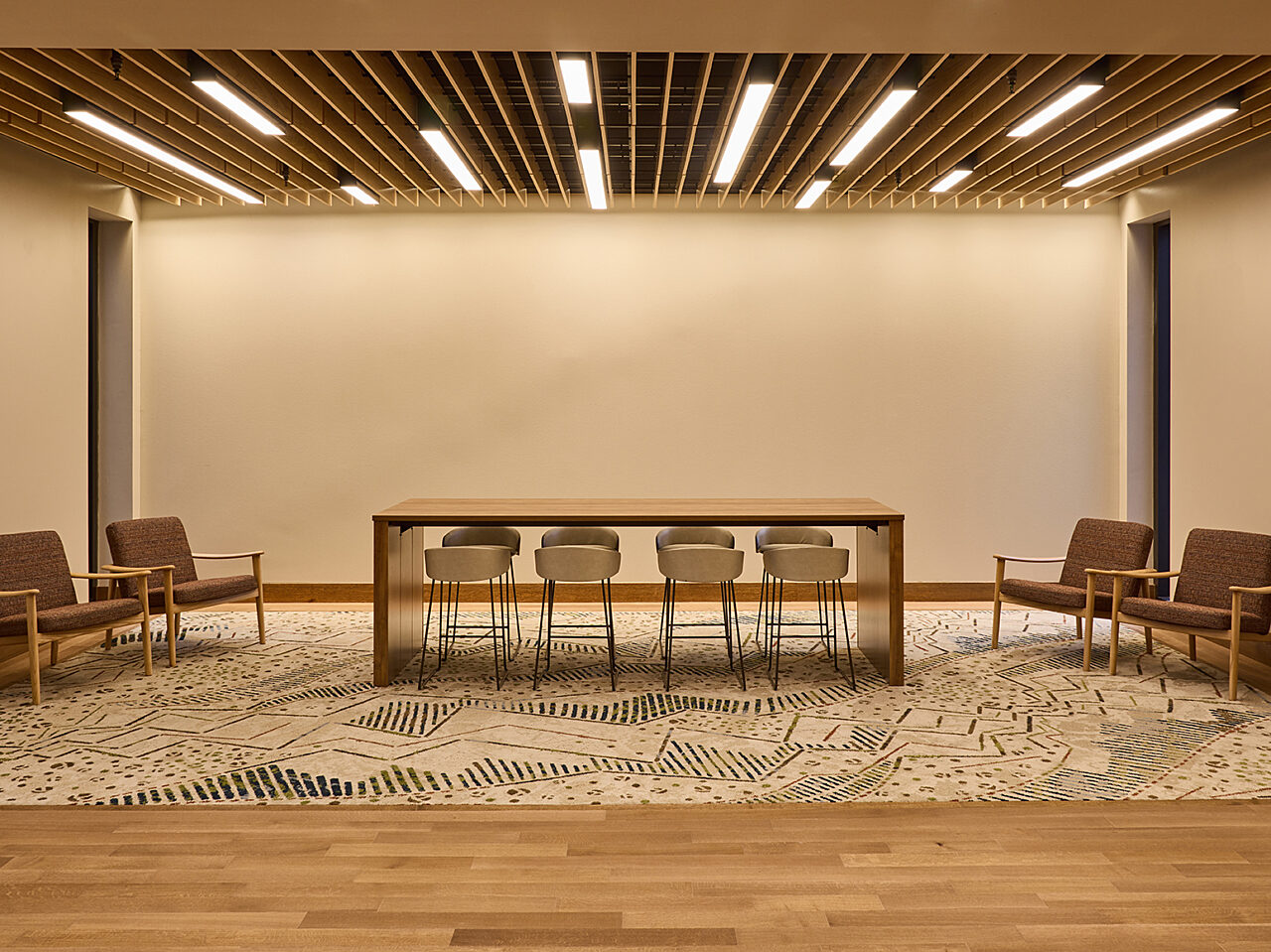 Collaboration area in hall with wood parsons table and low backed stools. Large patterned rug. Two lounge chairs flank each side of table. Slatted ceiling.