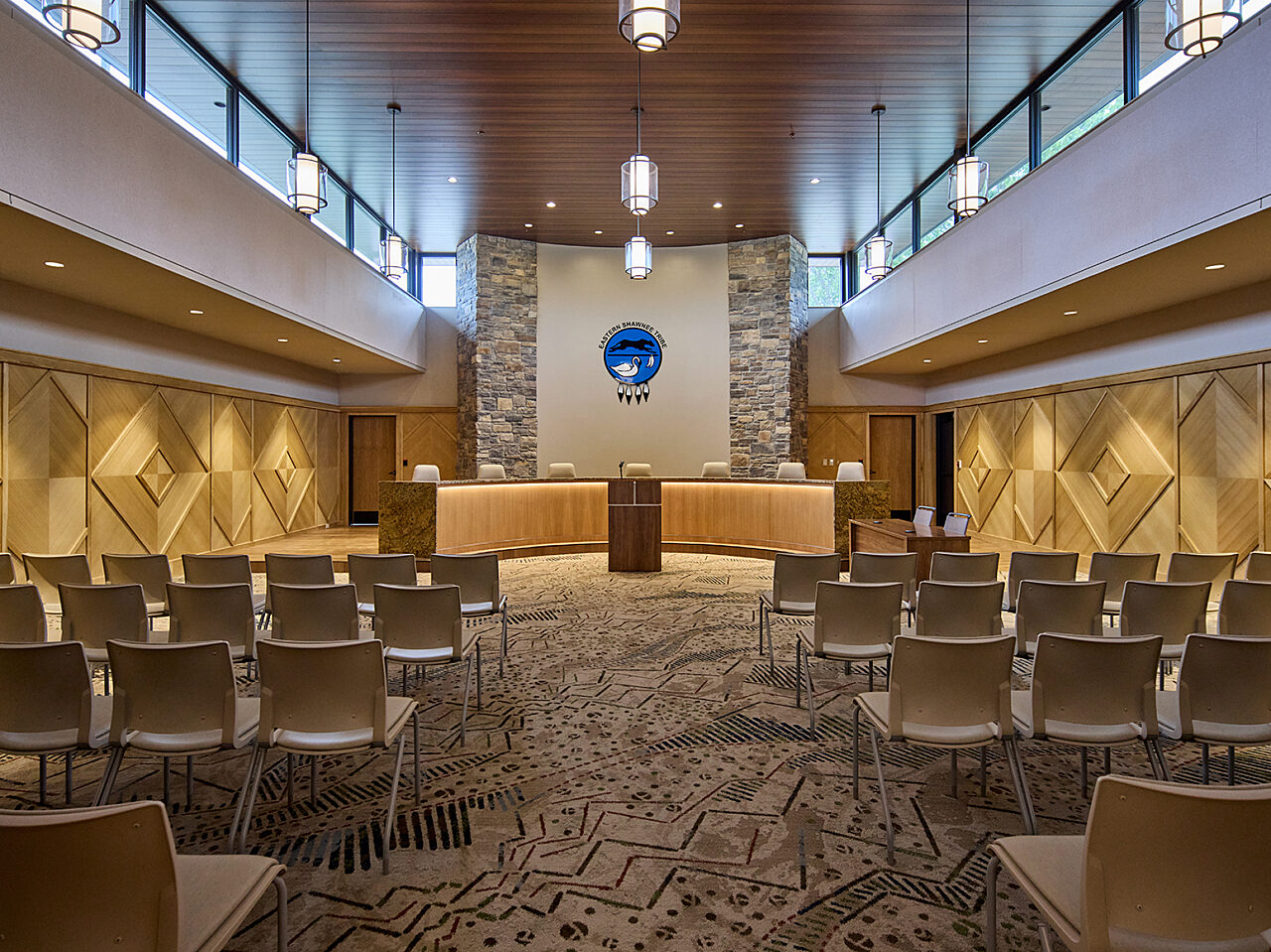 Large meeting hall with semi-circular desk on far end with Tribe logo on tall wall w/ stone. Mood lighting on wood walls. Chairs rows in foreground.