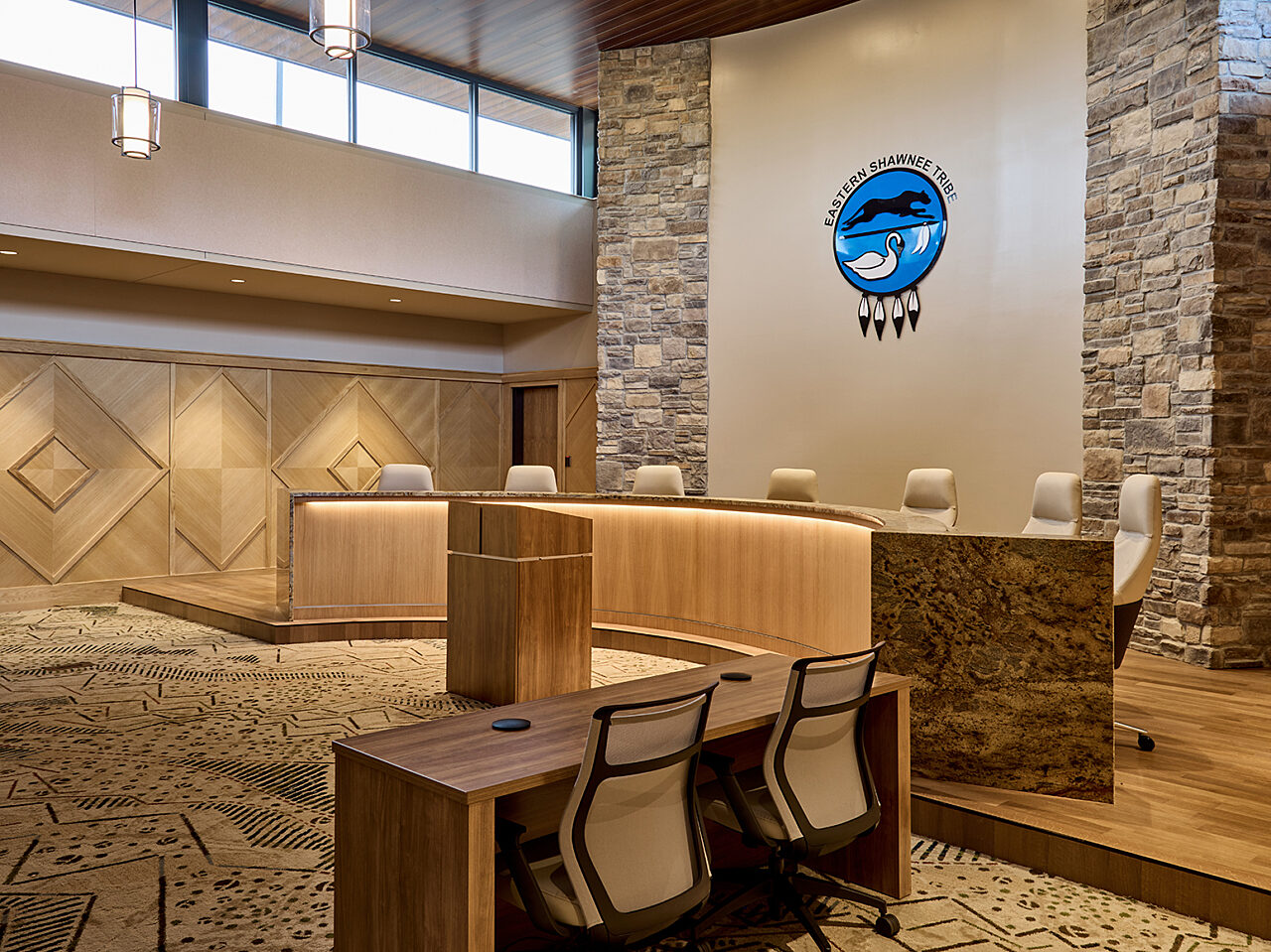 Meeting room with Eastern Shawnee Tribe logo above semi-circular granite/wood desk with 7 white leather chairs. Stone/wood walls and drop pendant lighting.