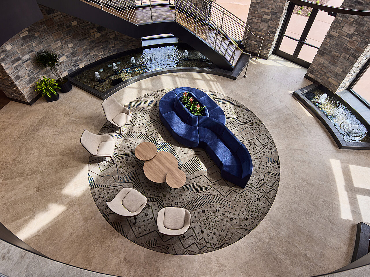 View looking down at seating area. Circular rug with a blue curved sofa, white lounge chairs and 3 nested coffee tables. Water features and rock walls.