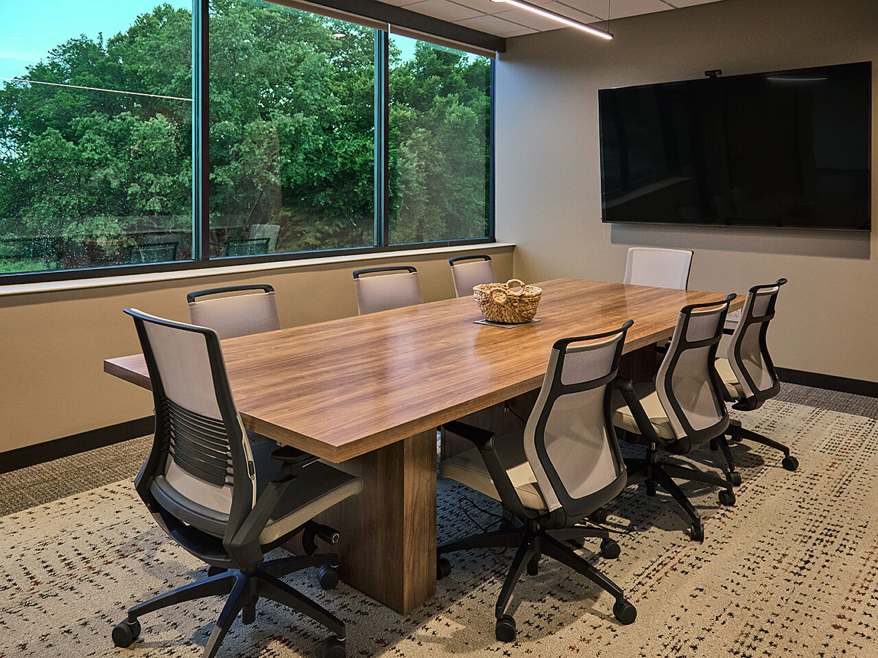 A conference room overlooking trees. Brown wooden table with 8 beige midback rolling task chairs. Mounted tv on one end of room.
