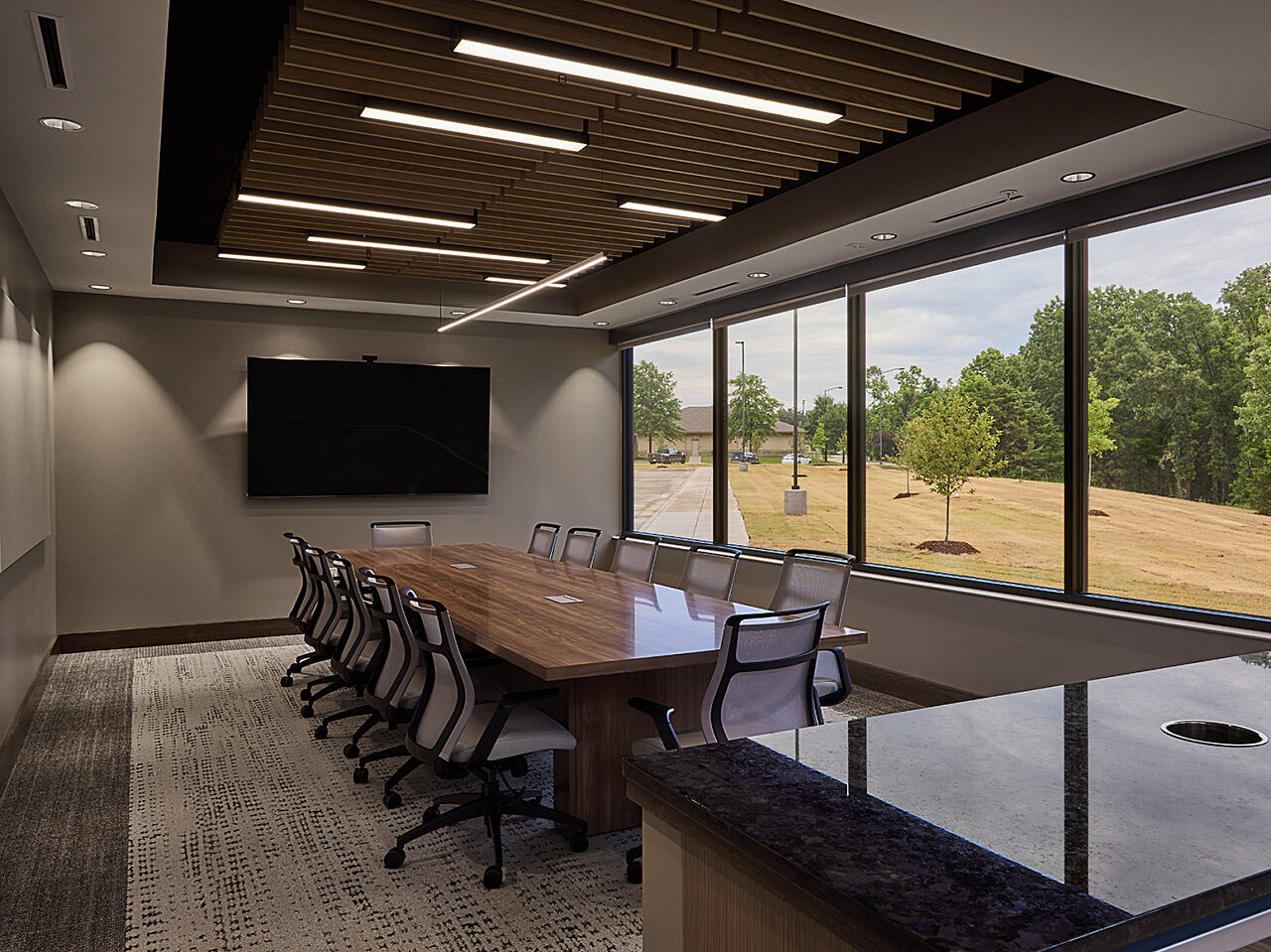 Executive conference room with 12 chairs around wood table, view of grass and trees outside. Mounted TV and easel board wall opposite wall of windows