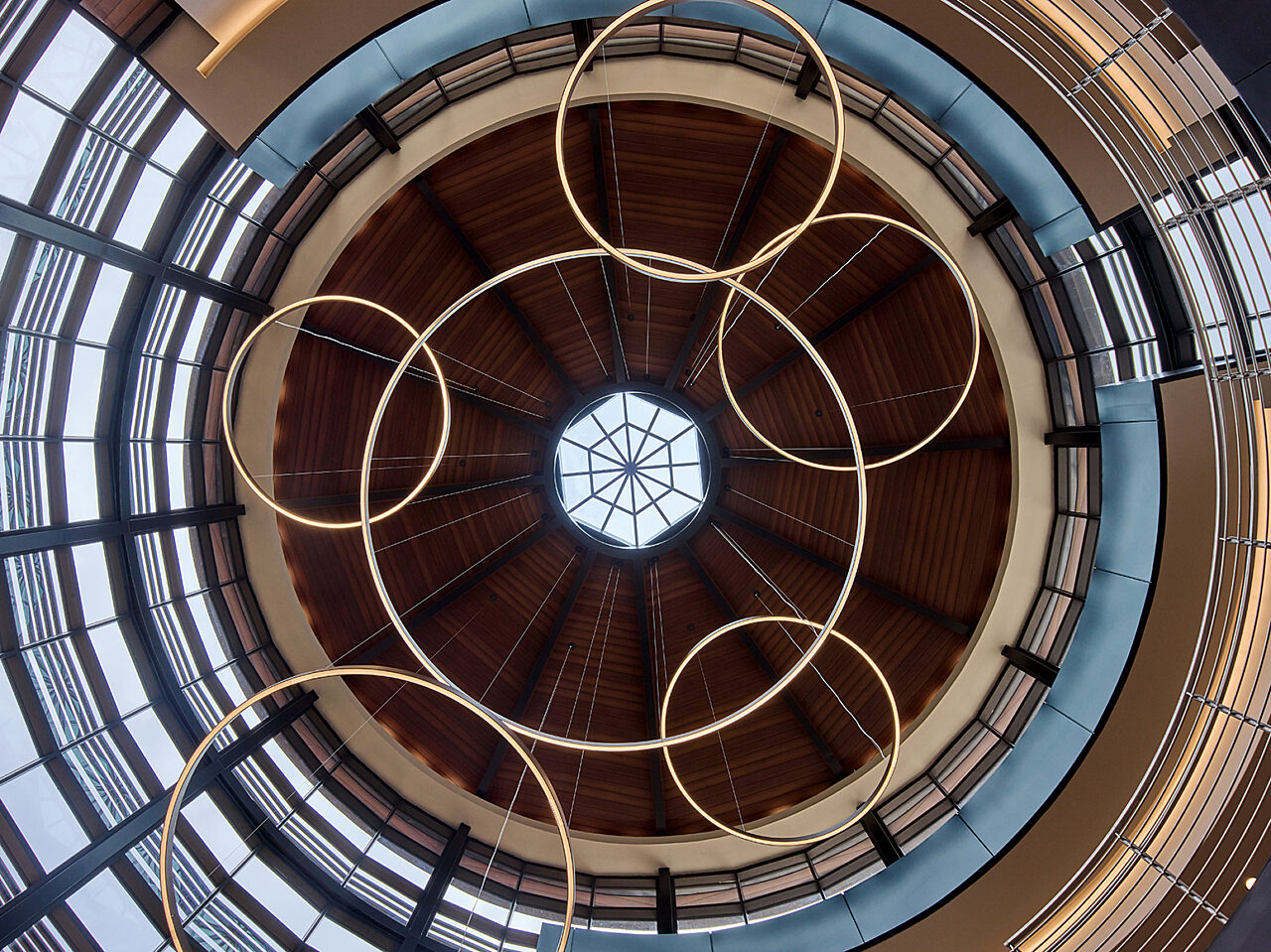 Ceiling view of cylindrical-shaped building. Some walls are wood, others are windows. Circular hanging lights form pattern around center hexagon skylight