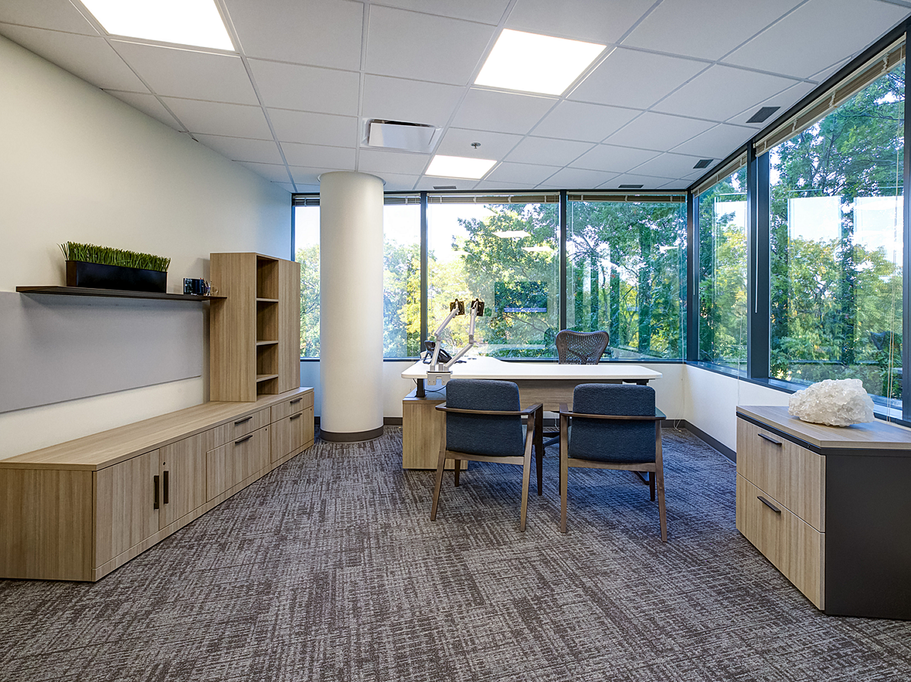 Corner executive office with large credenza, whiteboard and matching filing cabinet. Height adjustable desk with two blue/wood side chairs.