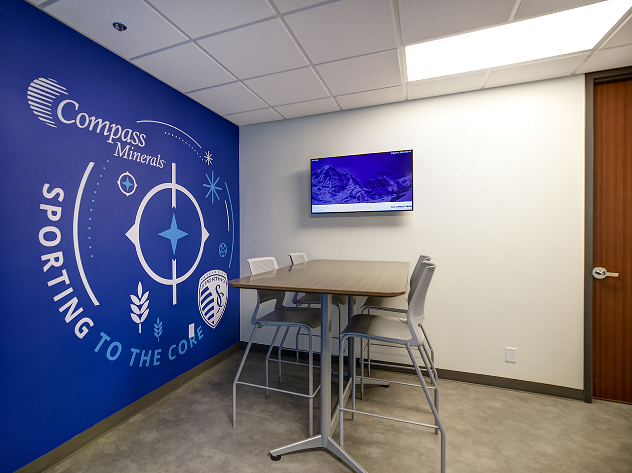 Compass Minerals touch-down area with blue Sporting KC wall featuring high top table and 4 stools with mounted tv