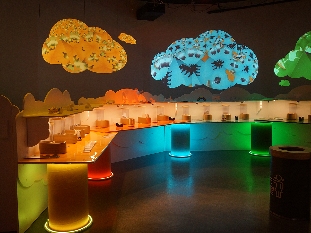 Hallmark Kaleidoscope art stations in a dimly lit room with light-backed colorful clouds and pillars.  A long desk with art supplies spans the room.