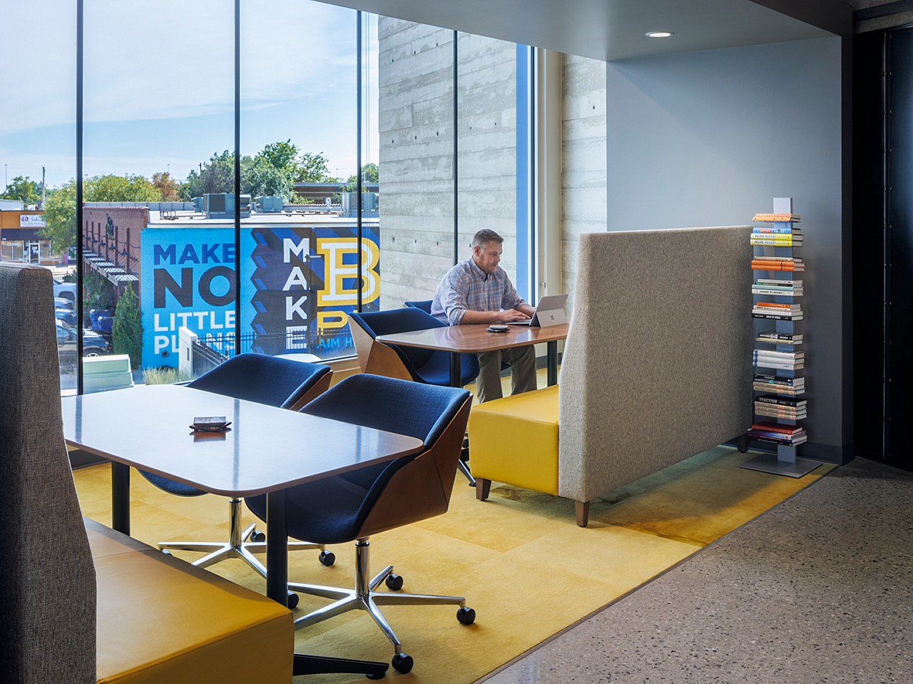 Hutton Construction-Yellow/Blue Focus area with yellow rug and yellow/grey banquette seating, tables, and blue rolling chairs. A man works on a laptop.