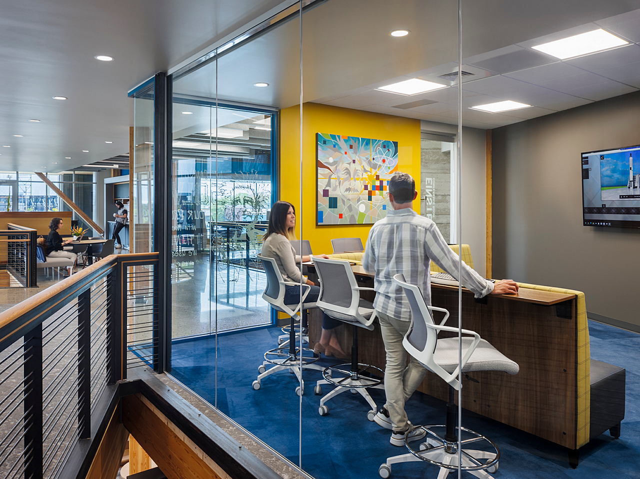 Hutton Construction-Colorful enclosed meeting area with modular seating with attached work counter and white rolling stools. Outside room are sitting areas