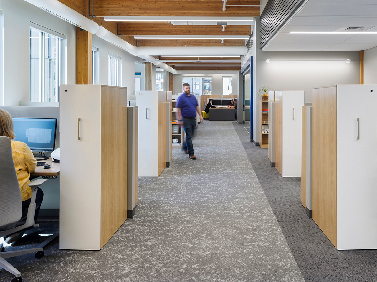 Hutton Construction corridor with workstations with lockers/storage on either side. A collaboration table and meeting are seen at end of hallway.