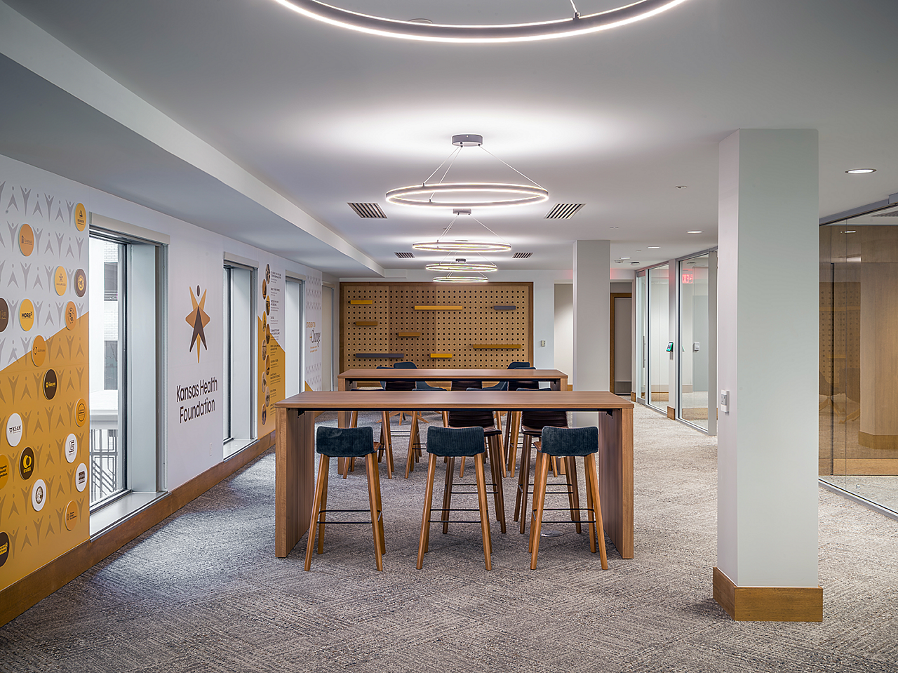 Collaborative area with 2 brown wood parsons tables and low back, upholstered stools. Kansas Health Foundation art on walls. Wood pegboard on far wall