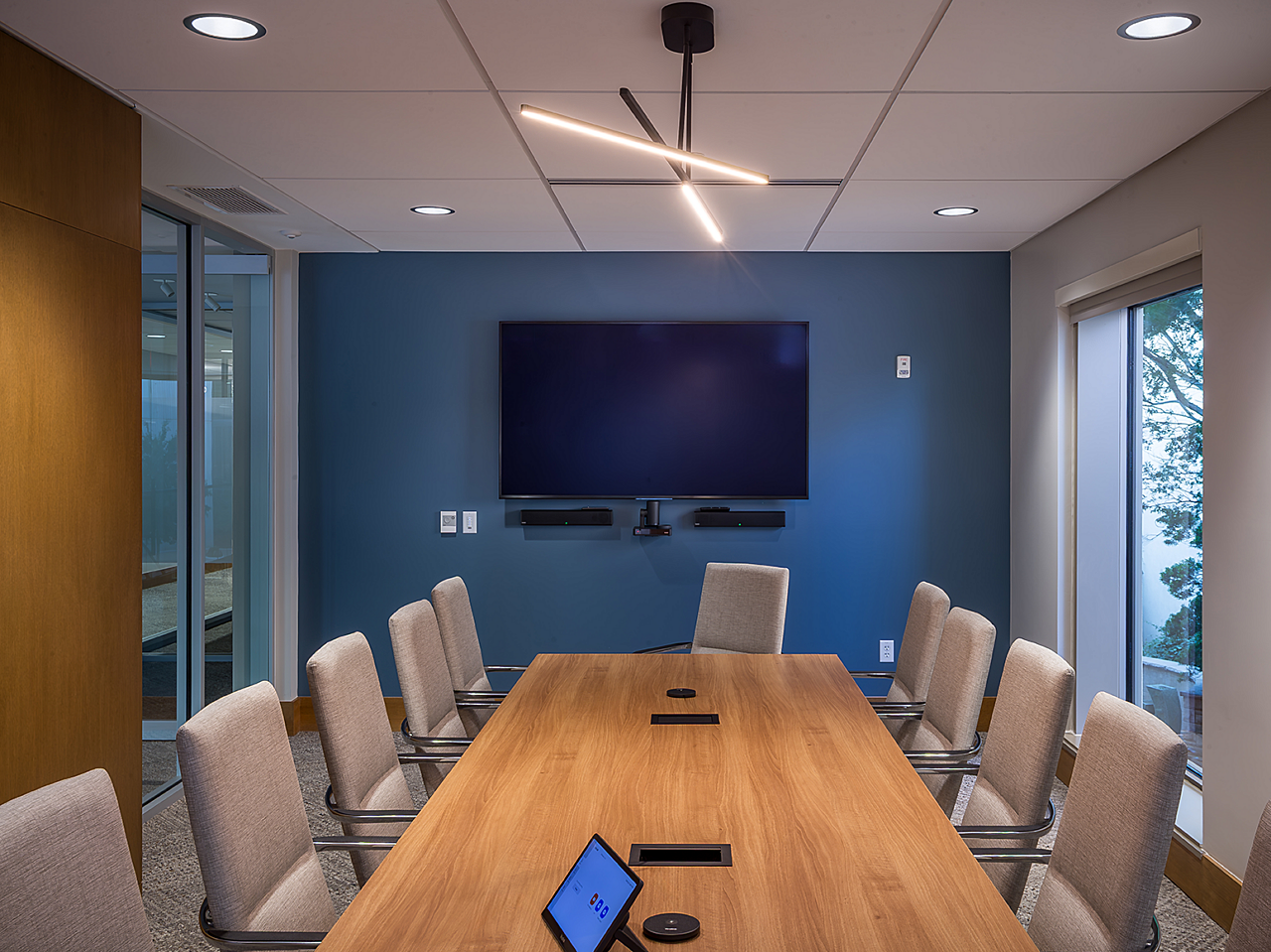 Conference room, blue wall with mounted TV, wooden conference table lined with upholstered high back chairs in a putty color. A modern, lamp hangs over