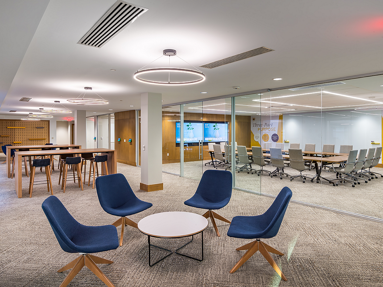 Conference room in background, lounge area in front with blue/wood lounge chairs and coffee table, and 2 parsons tables with blue upholstered stools behind