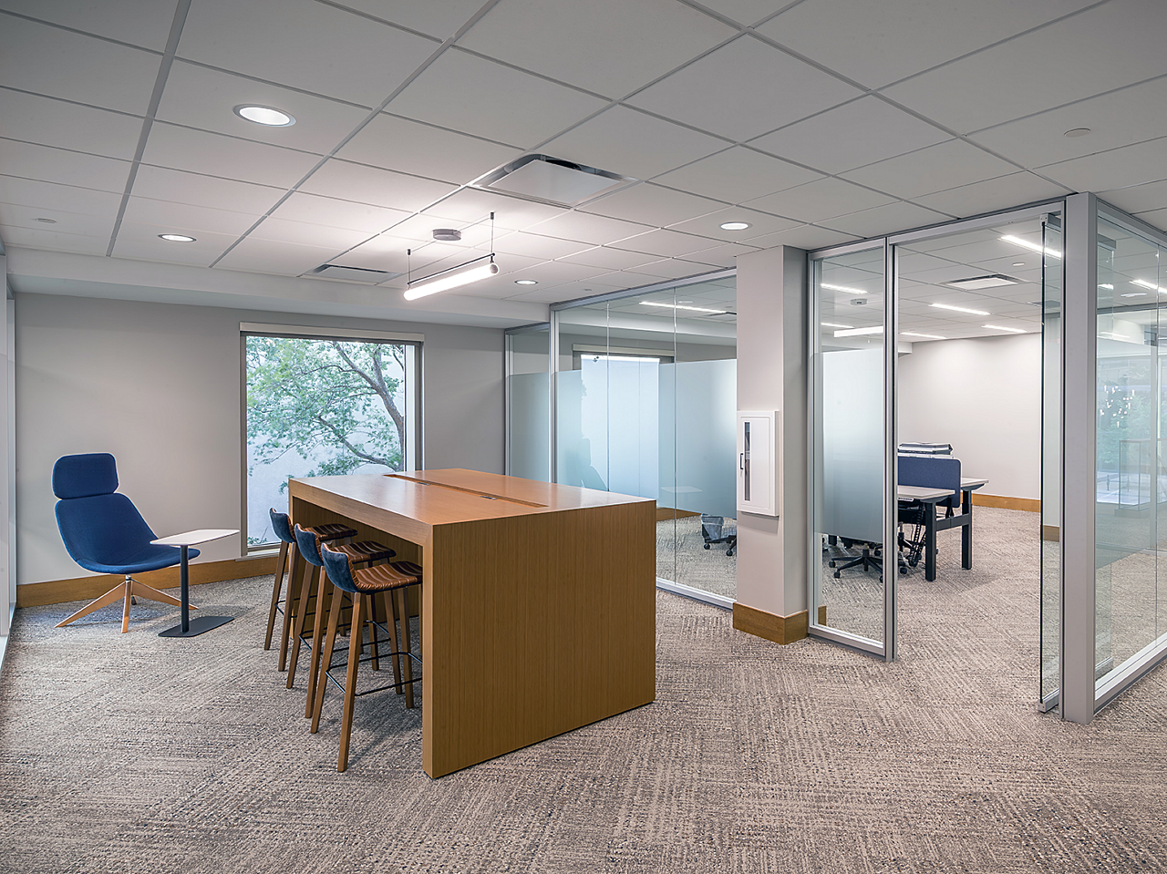 Collab area outside a glass wall office. Wide parsons table with 3 low-back stools with blue fabric on back. One high-back lounge chair with laptop table.