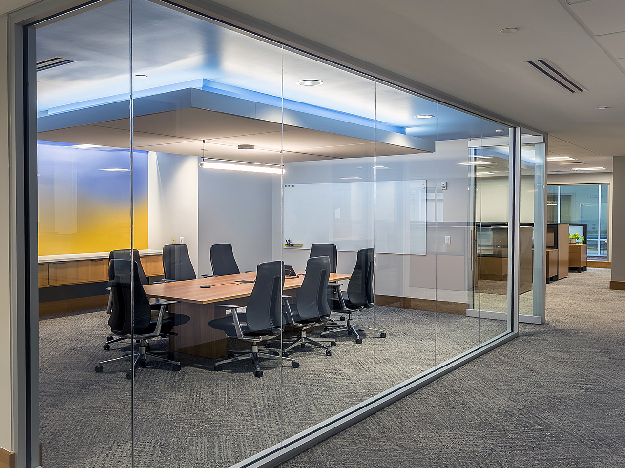 Glass wall conference room with wood table and black chairs. Yellow/blue gradient wall and one whiteboard wall. Dropped ceiling above table w/ blue border