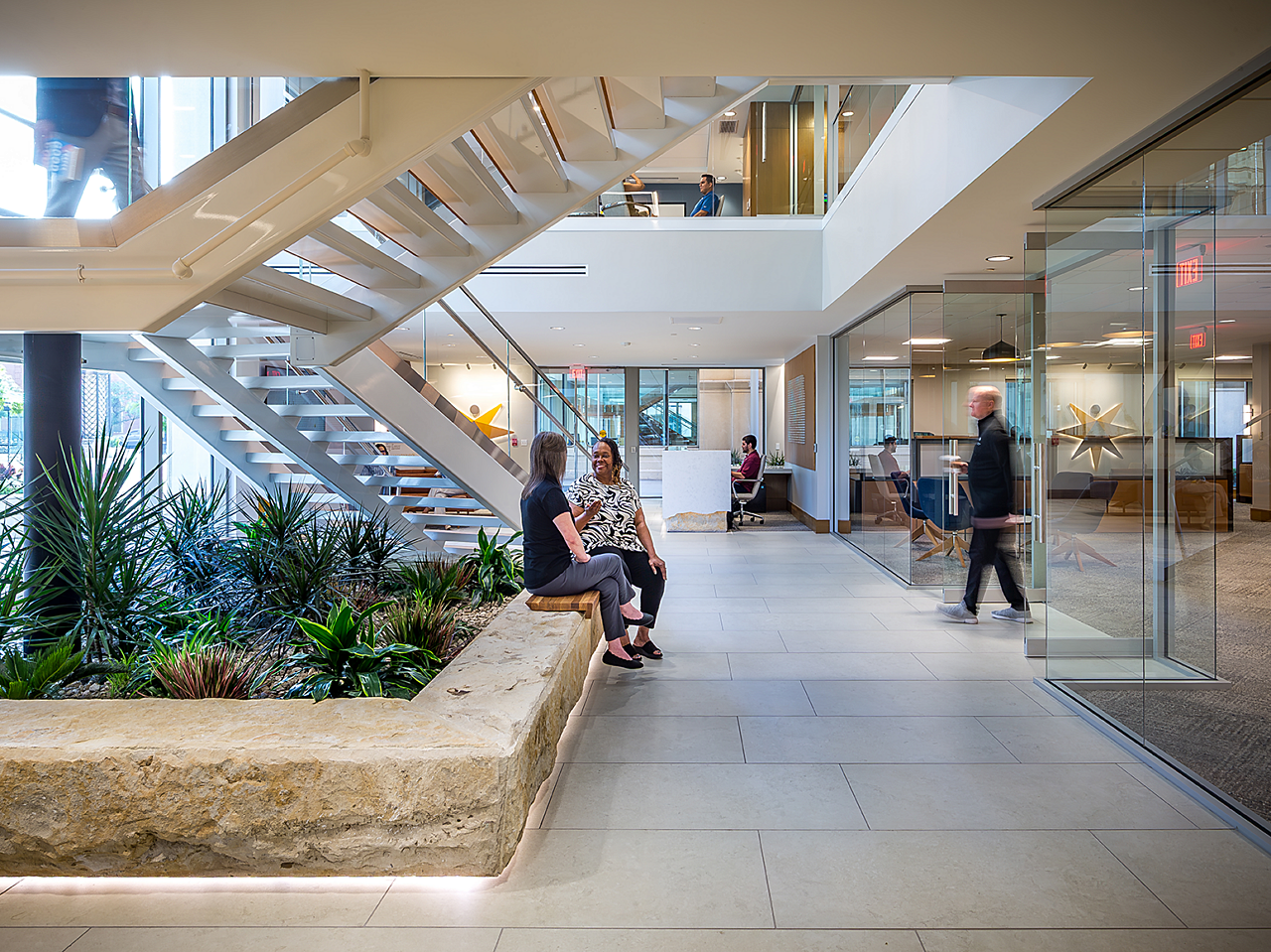Atrium area. Two women sit and chat on rock walls under staircase. Reception area in background. Man walking out of workspace with glass doors