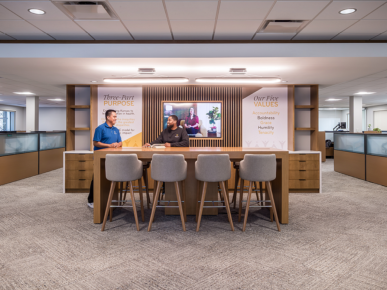 Huddle Space/Collab area. Two mean stand at table. Back cabinets and wall with mounted TV and company mission/verbiage. Workstations to either side of them
