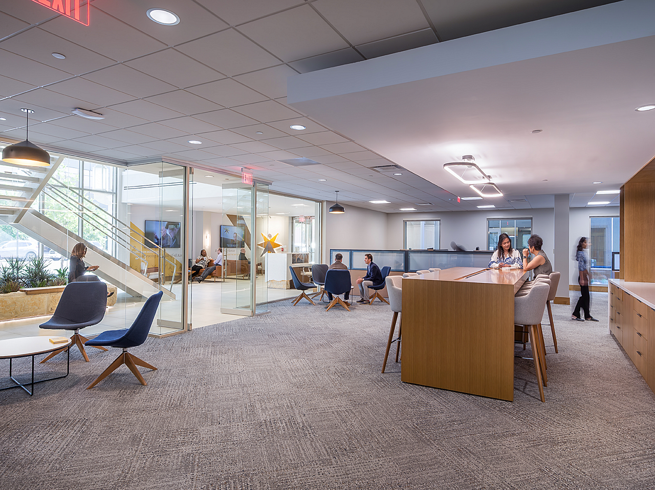 Two lounge sitting areas to each side of the doorway, near walls of windows. Two women chat while sitting on stools at a collaborative parsons table.