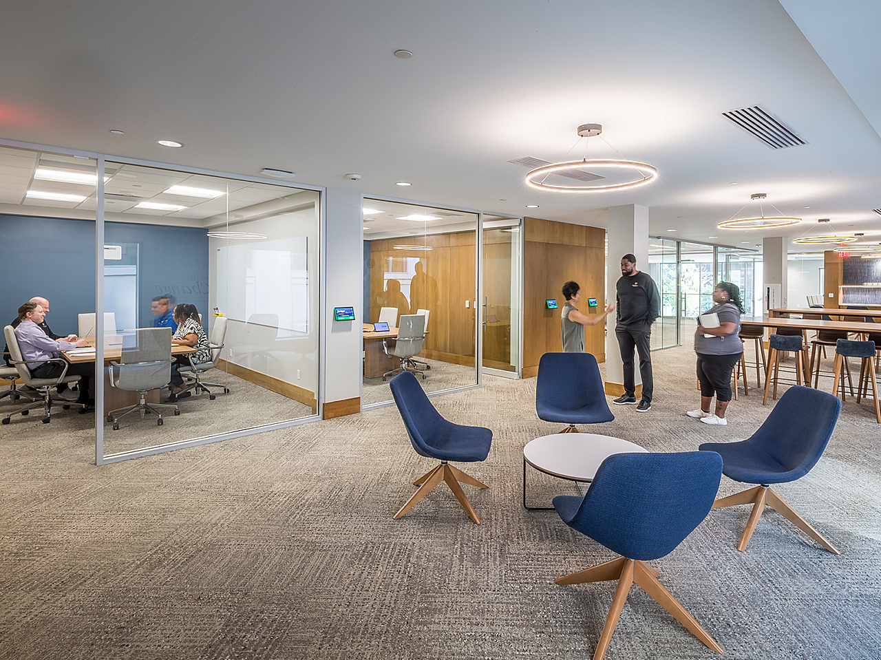 Open office with multiple glass-walled conference rooms, 4 lounge chairs and a round coffee table in front, high meeting tables with blue stools on right