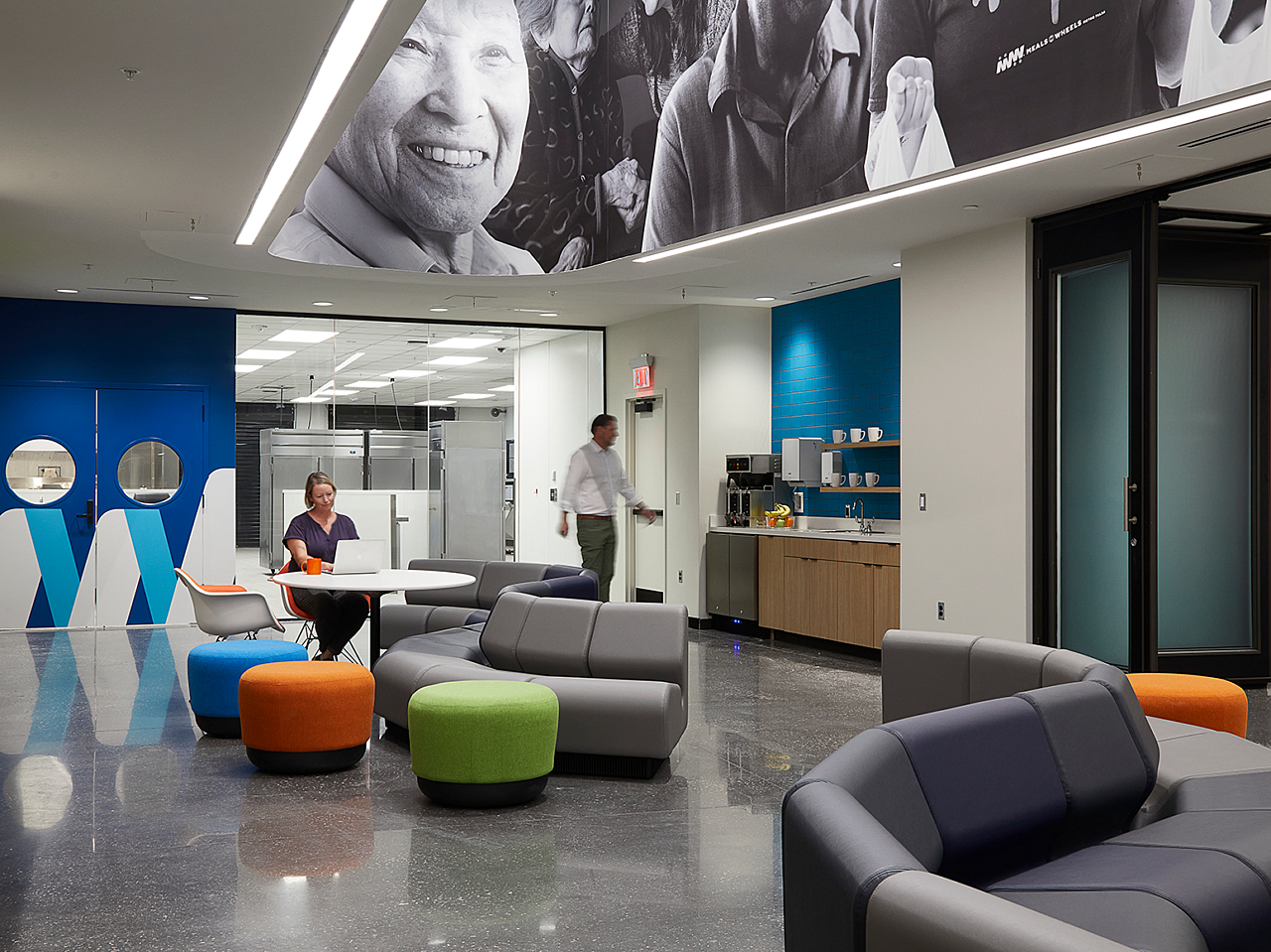 Hallway workspace/coffee area. Woman sits at table with laptop. Wavy-shaped grey modular sofas with bright blue, orange and lime pouf ottomans around them