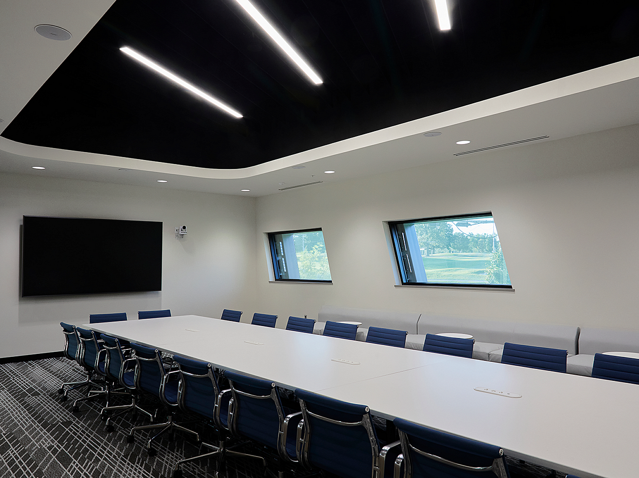 Conference room with long white table and blue Eames office chairs. Two cut-out windows along walll and a large mounted tv on adjining wall.