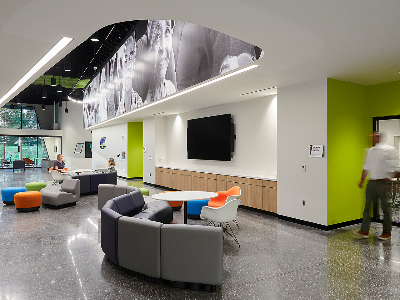 Tulsa Meals on Wheels-Hallway with lime/white paint and b&w people mural on top. Wavy-shaped grey couch with colorful pouf ottomans and lounge chair.