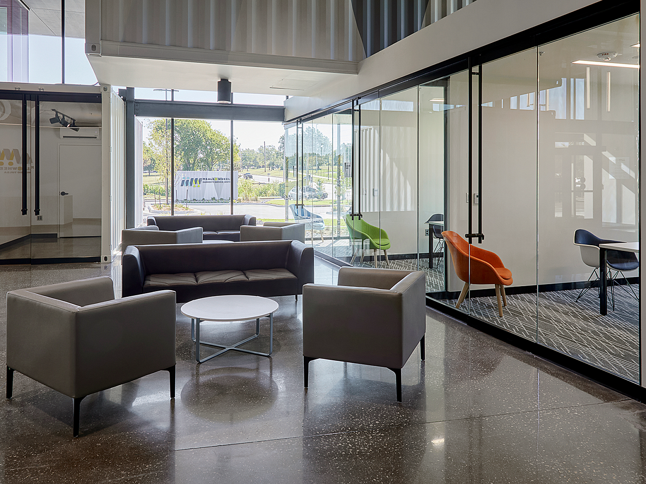 Two groups of lounge seating with grey couch and chairs around round coffee table.  Glass-walled offices along one wall with colorful side chairs.