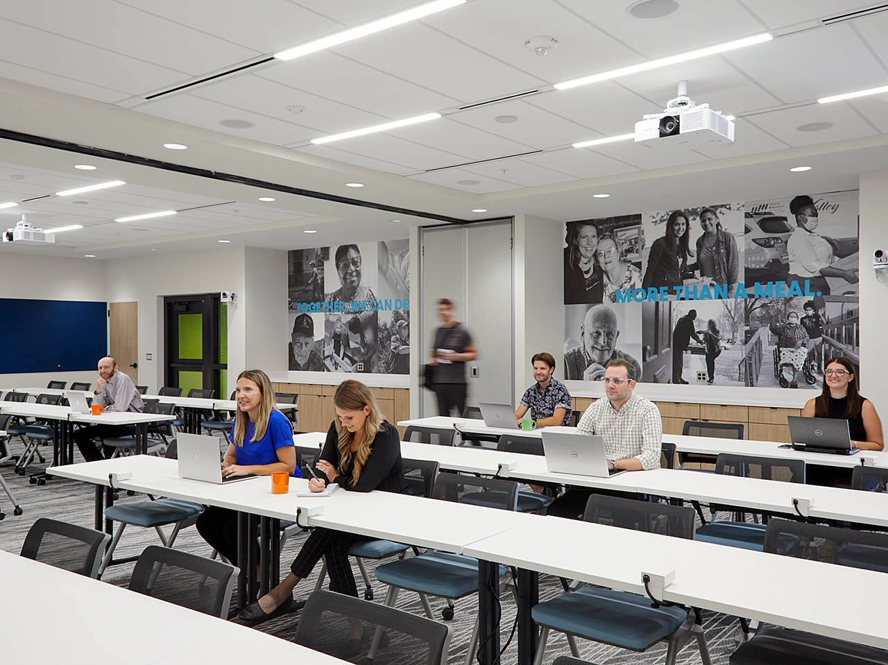 Large training room with long tables and black chairs. Employees sit with laptops. Back wall has black and white photos of people Meals on Wheels helps