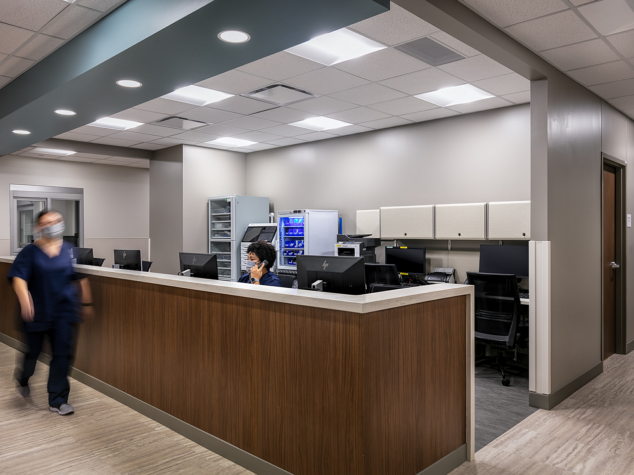Saint Luke's South - Nurses's station and seating. A masked nurse sits at the counter using the phone, while another walks by on her way to an appointment.