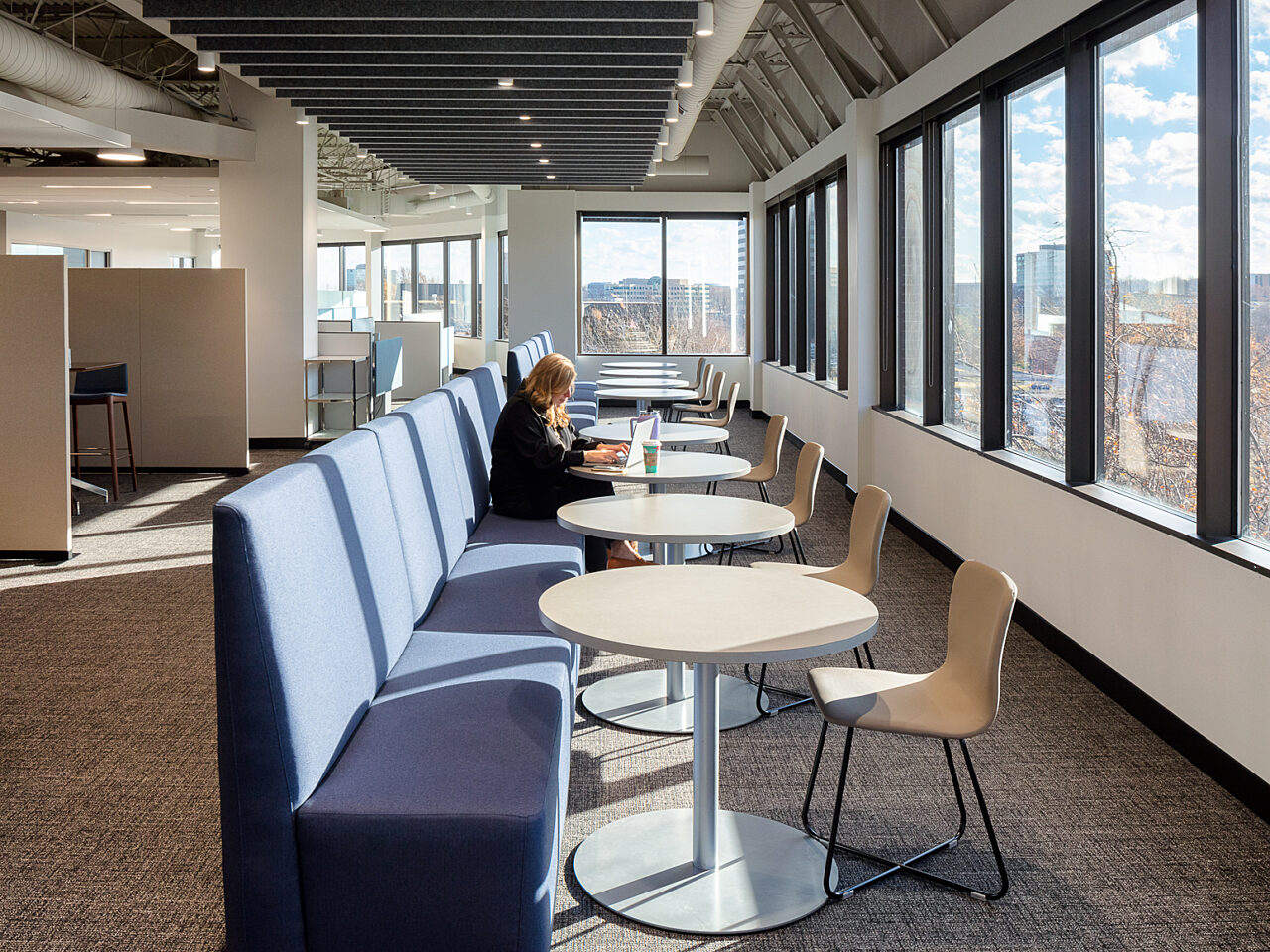 Two sets of long blue banquet seats with steel/wood cafe tables and beige stools along a wall of windows. A lone woman works on a laptop at a table