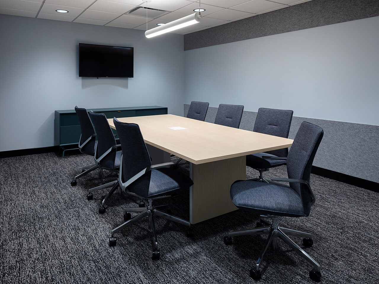 A small conference room with wooden table, upholstered rolling chairs in dark grey. A long white board covers the wall, along with a large screen tv .