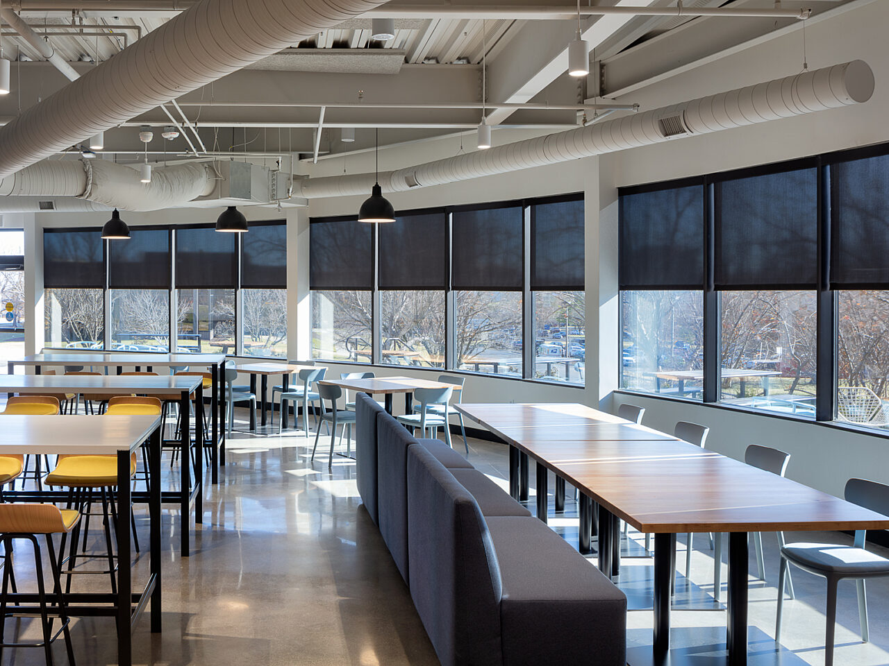 Window-lined cafeteria with multiple seating options: banquette seating, high-top tables and stools and rectangular dining tables with chairs