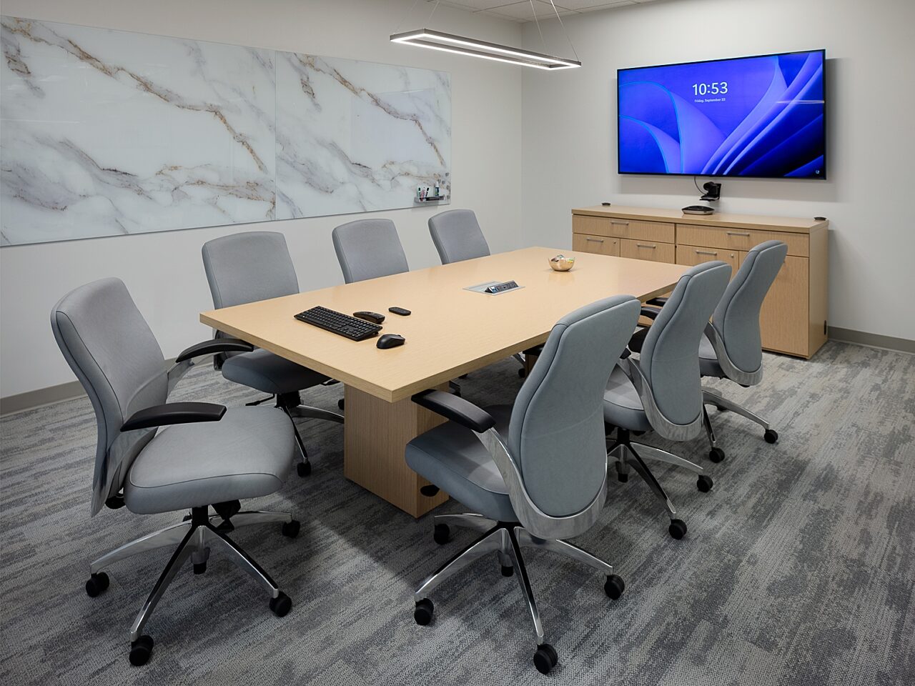 Conference room with light wooden table and credenza. Grey, highback chairs with casters a marbled glassboard on the wall that resembles art.