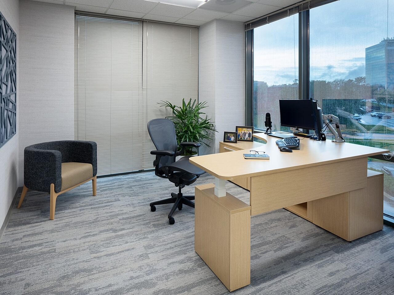 Executive office featuring a sleek, height-adjustable wooden corner desk,  Acoustical art on wall, and decorative u-shaped chair in the corner in black/tan