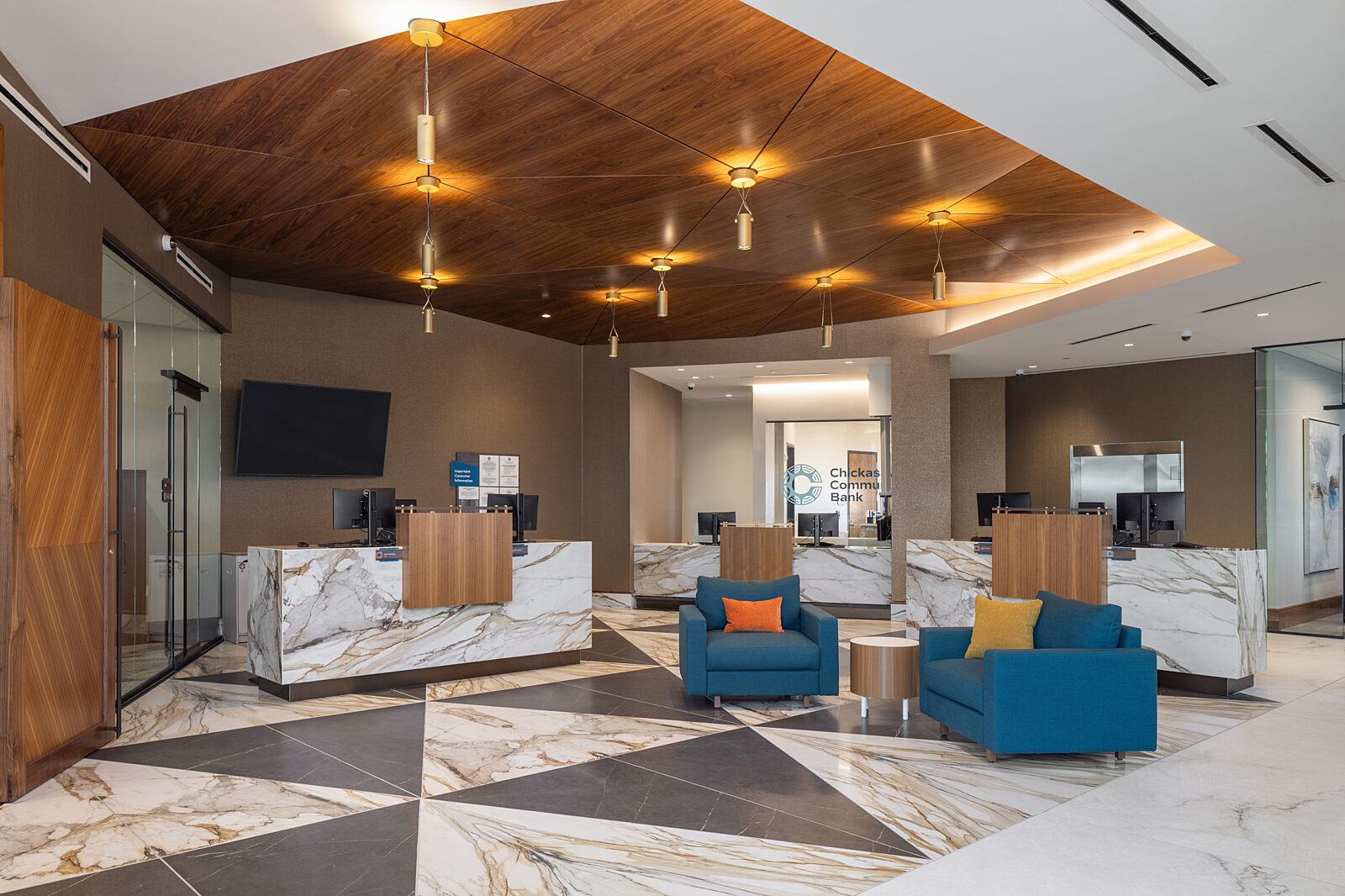 Bank lobby with marble flooring and counters, wood accents on doors, desks and as accent in ceiling. Blue lounge chair seating with pillows and side table