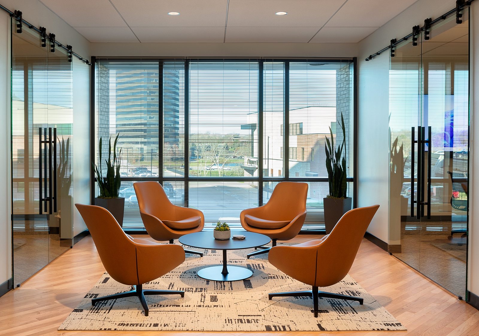 Lounge area between two conference rooms with sliding glass doors. Four tan, leather chairs and round coffee table. Tall plants and windows behind
