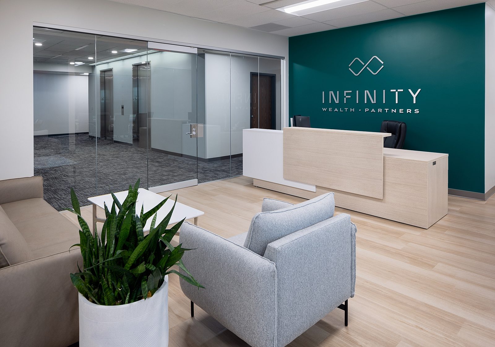 Infinity Wealth Partners in 3D silver letters on teal wall. Light wood/white accent reception desk, beige leather sofa, coffee table, grey chair, plant