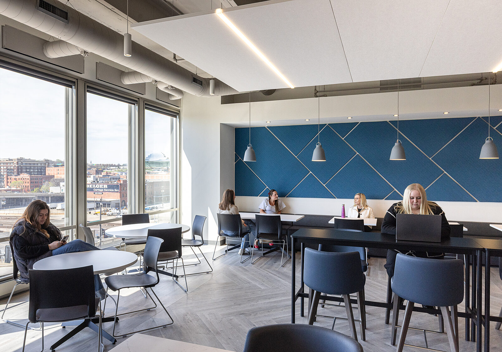 Cafe with five employees in multiple seating types including cafe, banquette and high-top collaboration table.  Blue wall on far w/ with grey pendant lamps