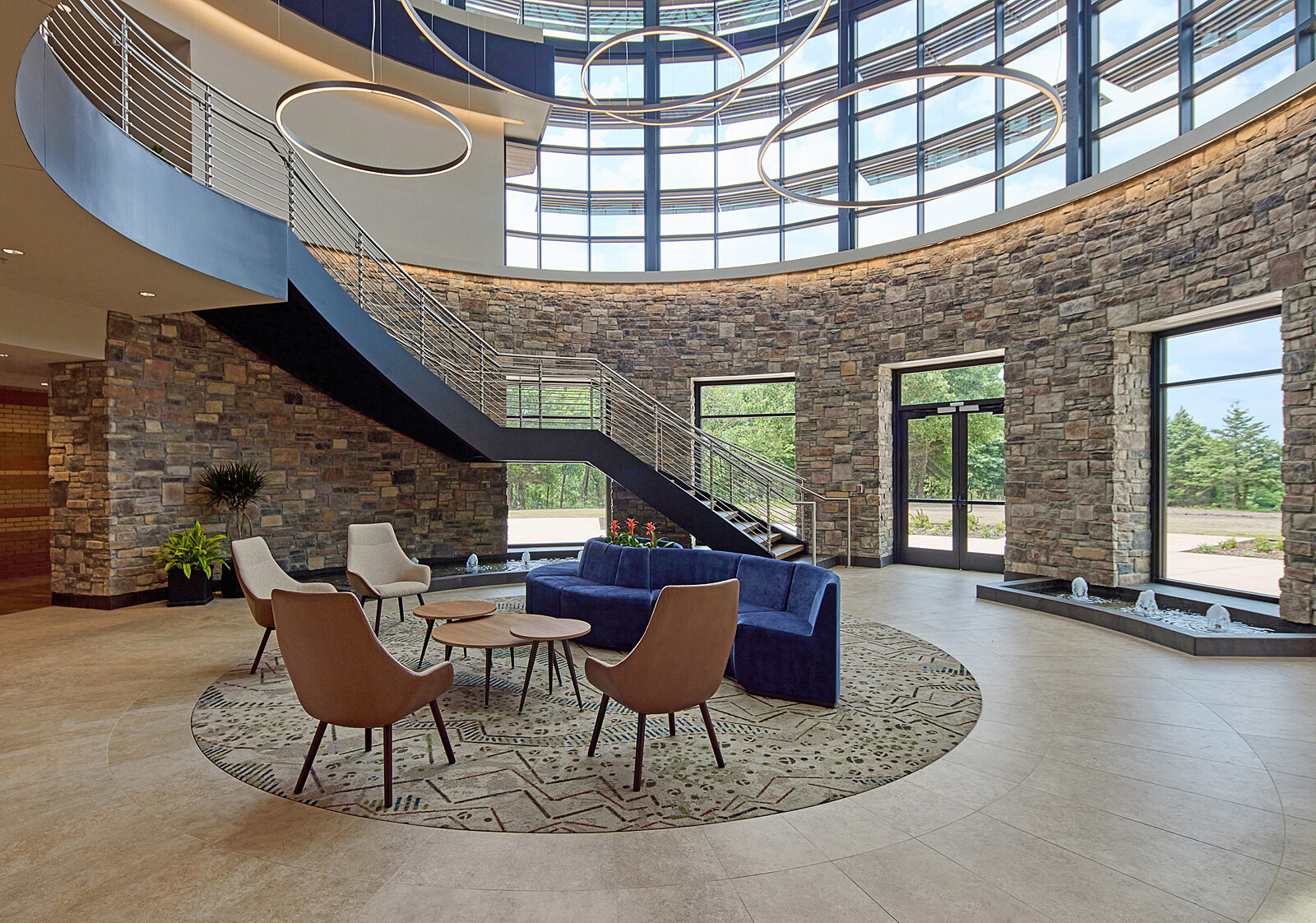 A cylindrical-shaped room with stone walls and windows. Found rug in center with lounge seating (blue couch & wood lounge chairs and coffee tables).