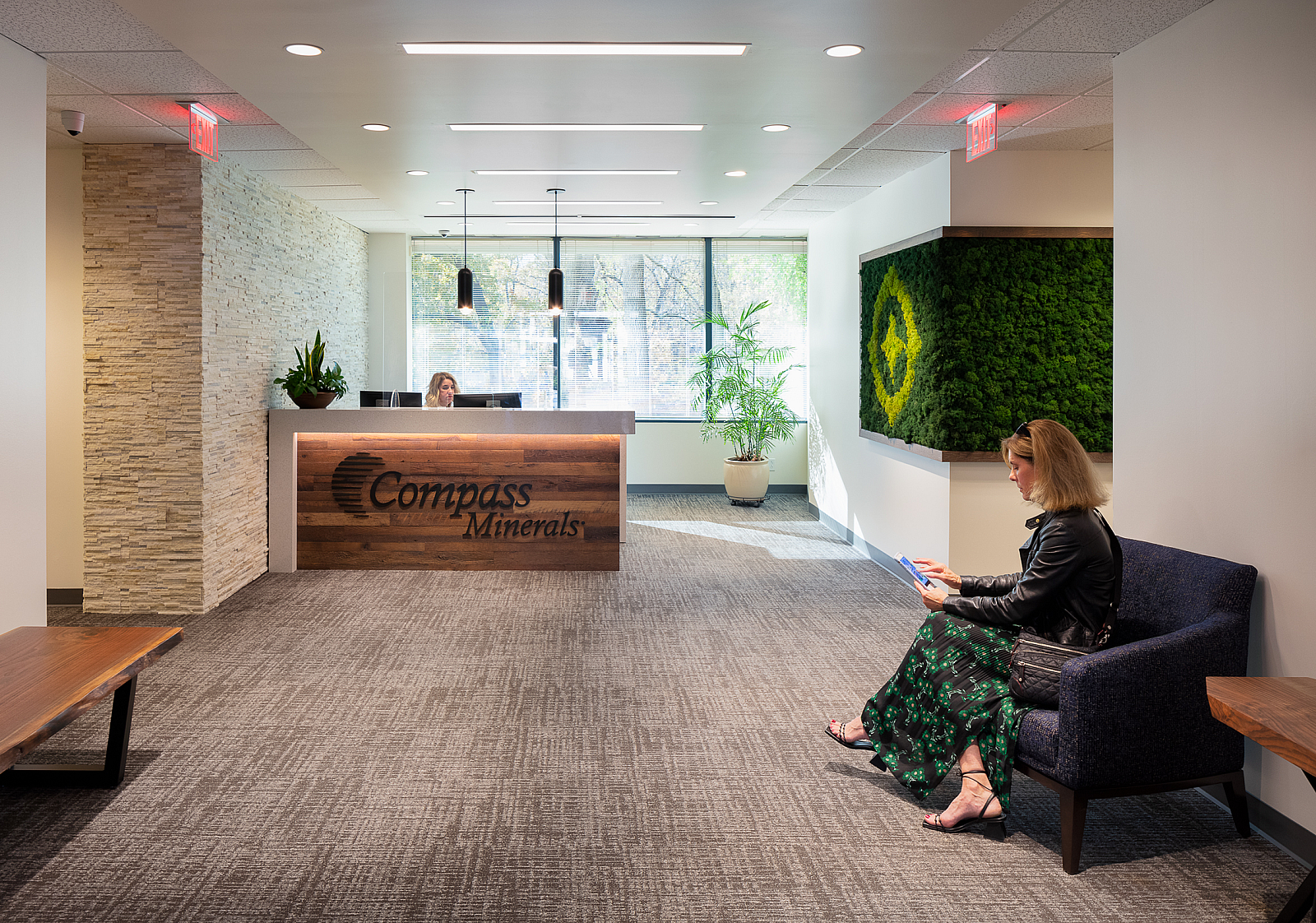 Compass Minerals Reception -tiled wall and moss feature. Receptionist sits and desk and woman in green/black dress and leather jacket sits in a blue chair.