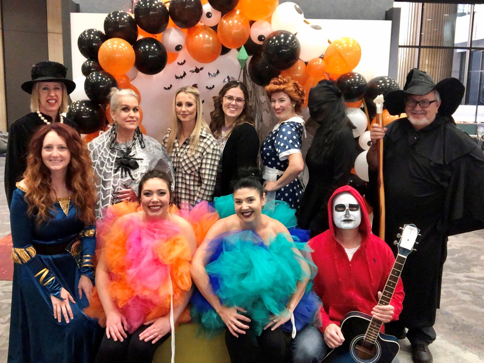 Employees dressed in Halloween costumes, standing in front of an orange and black balloon arch.