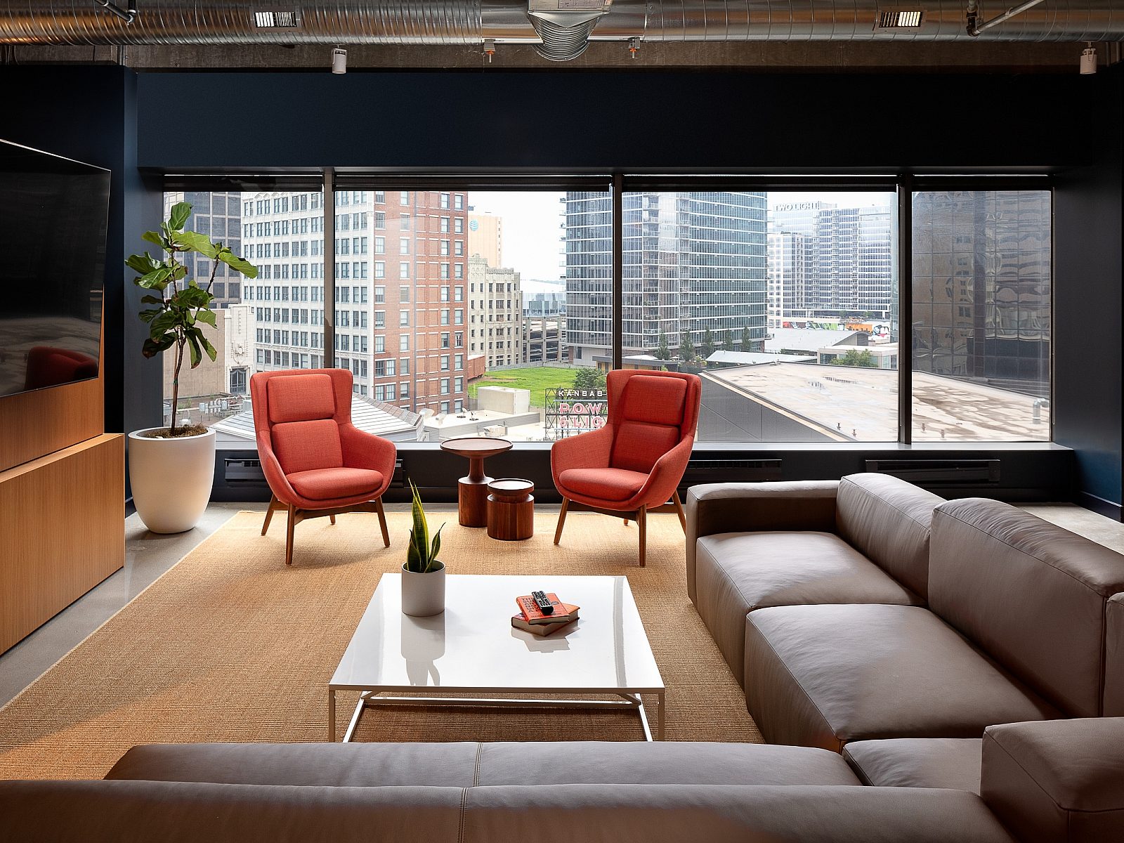PayIt lounge with view of city buildings. Two orange high-back lounge chairs, a tv, and L-shaped leather sofa, coffee table and industrial ceiling.