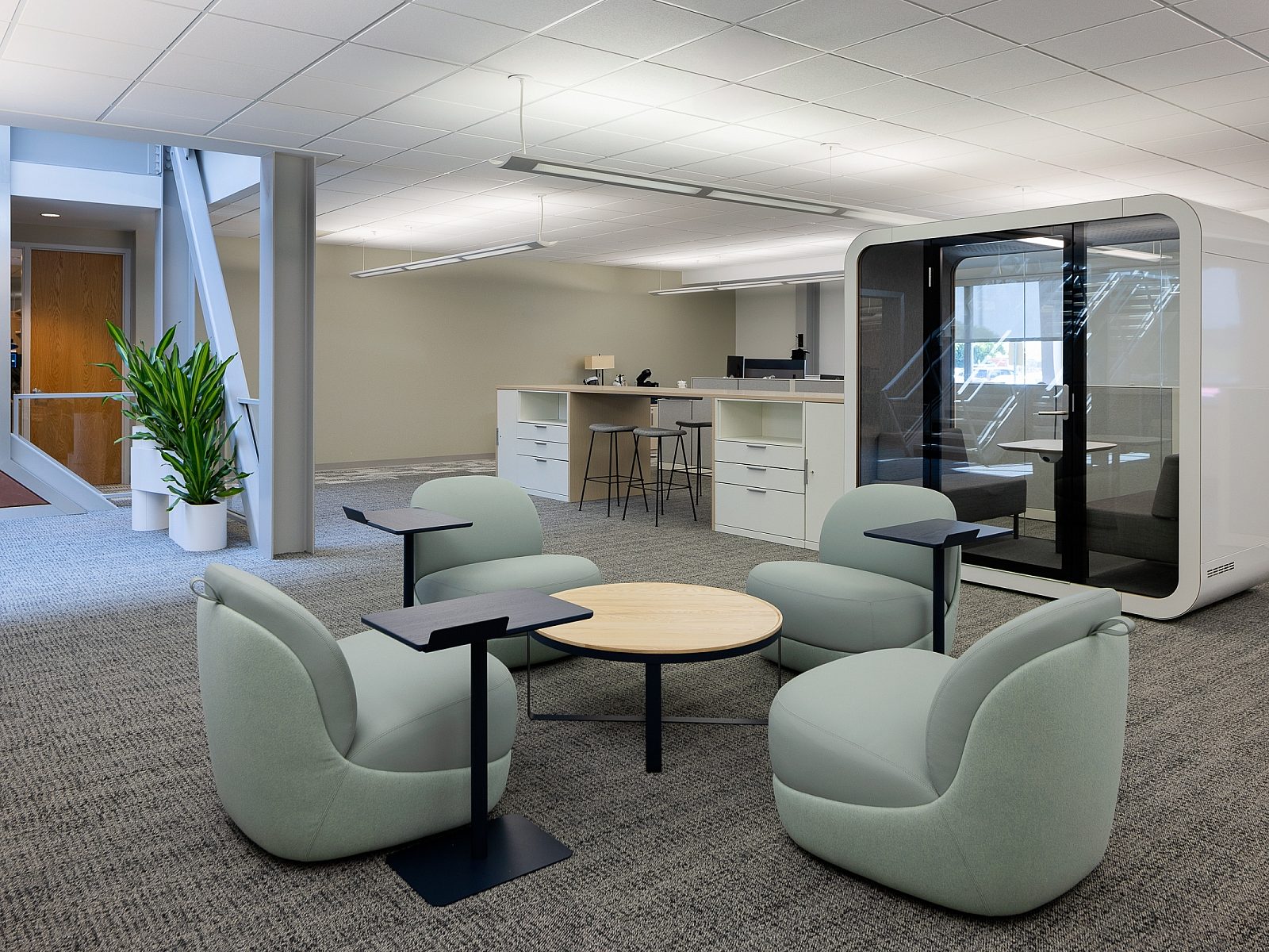 Bluescope - Open collab area with four pale blue armless lounge chairs around a tall coffee table with computer stands, Large Framery pod in background.