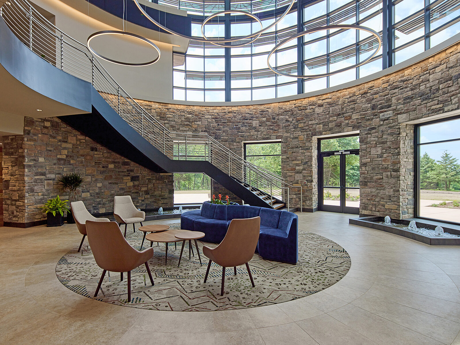 A cylindrical-shaped room with stone walls and windows. Found rug in center with lounge seating (blue couch & wood lounge chairs and coffee tables).