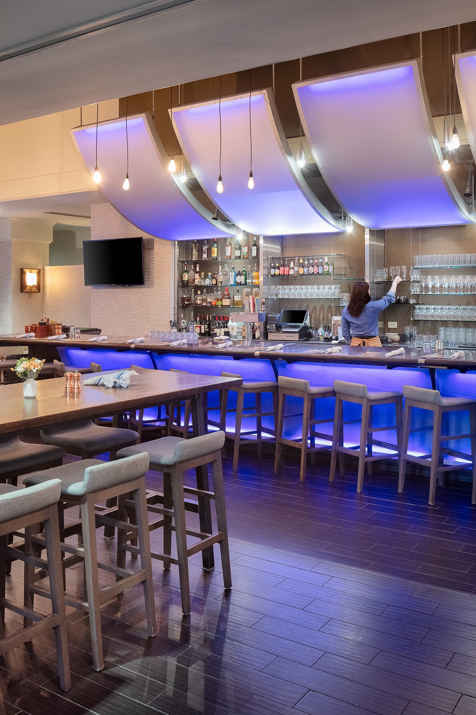 Hotel bar and rectangle bar tables with tall stools with upholstered seats and short backs. Bar lighting casts a blue shimmer. Single lightbulb ropes hang